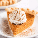 A slice of Classic Pumpkin Pie topped with whipped cream and dusted with cinnamon, served on a white plate.