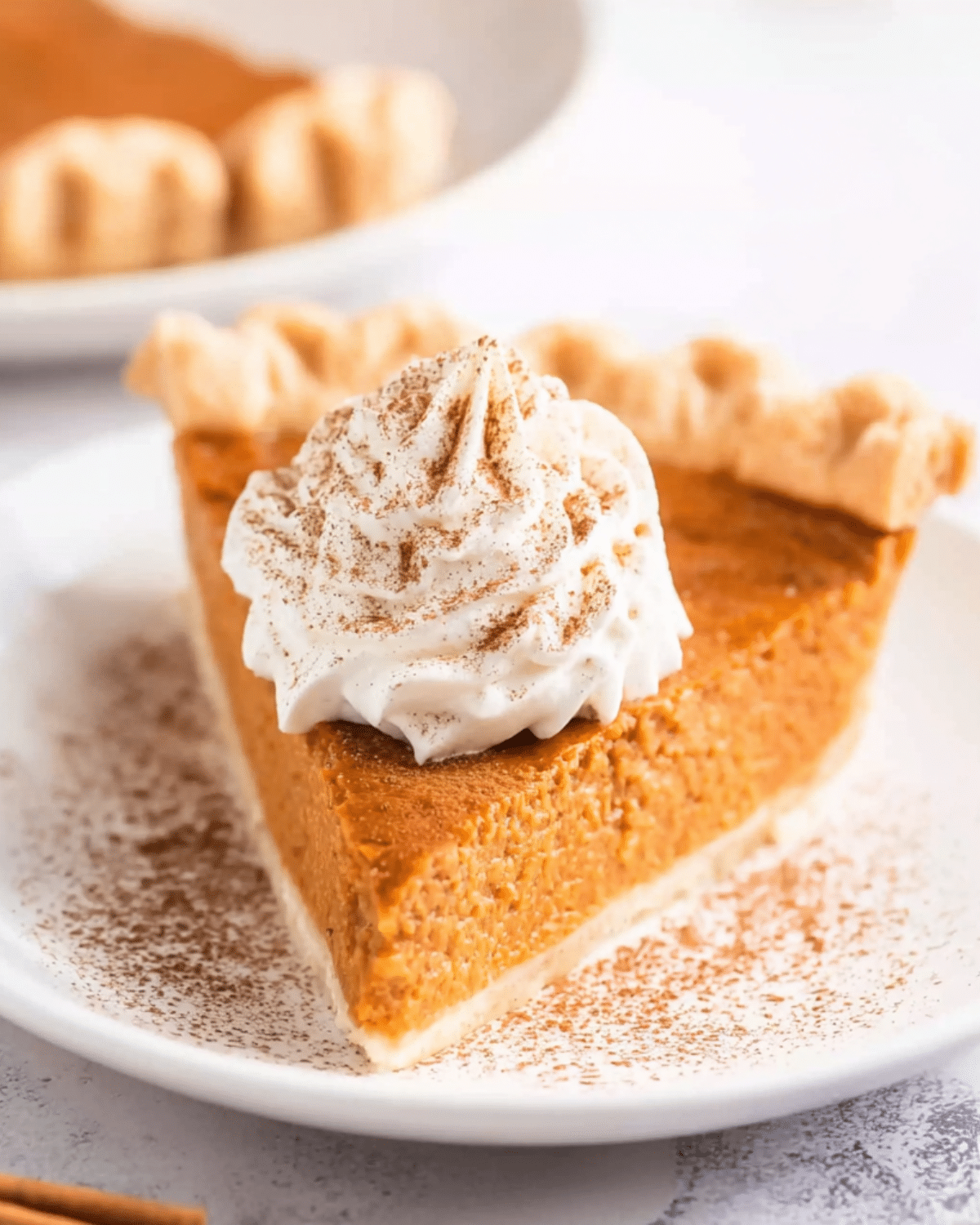 A slice of Classic Pumpkin Pie topped with whipped cream and dusted with cinnamon, served on a white plate.