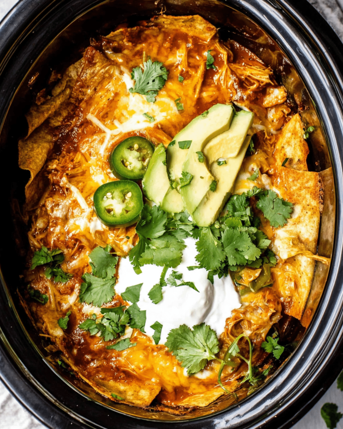 A crockpot filled with chicken enchilada casserole, topped with melted cheese, sour cream, avocado slices, jalapeños, and fresh cilantro.