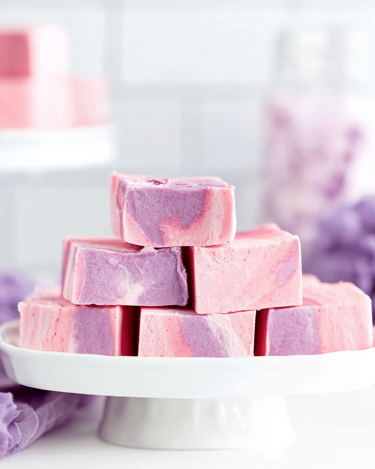 Stacked pieces of dreamy cotton candy fudge with pink and purple swirls displayed on a white cake stand.
