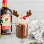 Festive Drunken Rudolph cocktail topped with whipped cream, chocolate antlers, and a cherry nose in a clear glass mug.