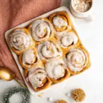 Festive cinnamon rolls topped with creamy icing in a white baking dish, surrounded by holiday decorations and a warm drink.