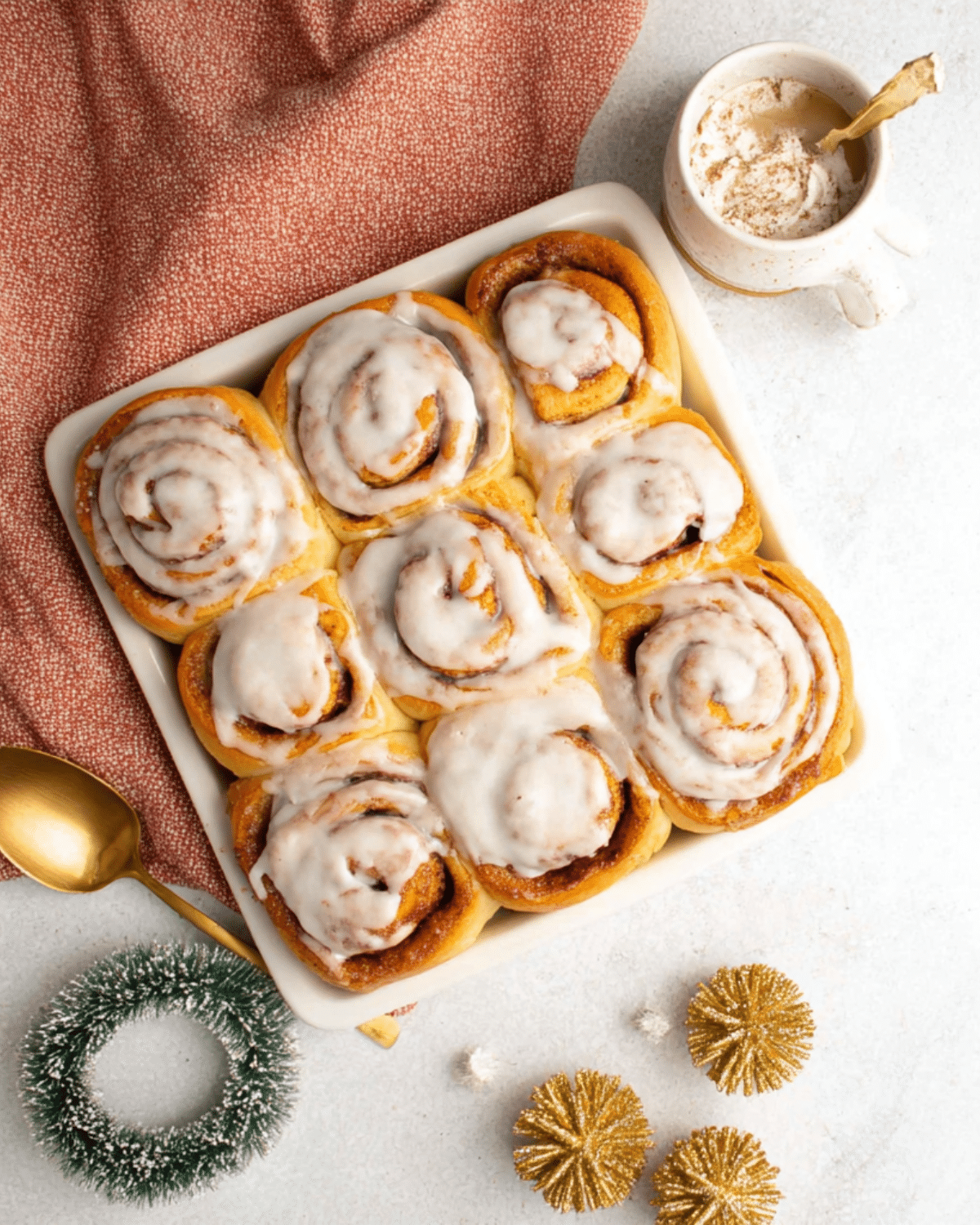 Festive cinnamon rolls topped with creamy icing in a white baking dish, surrounded by holiday decorations and a warm drink.