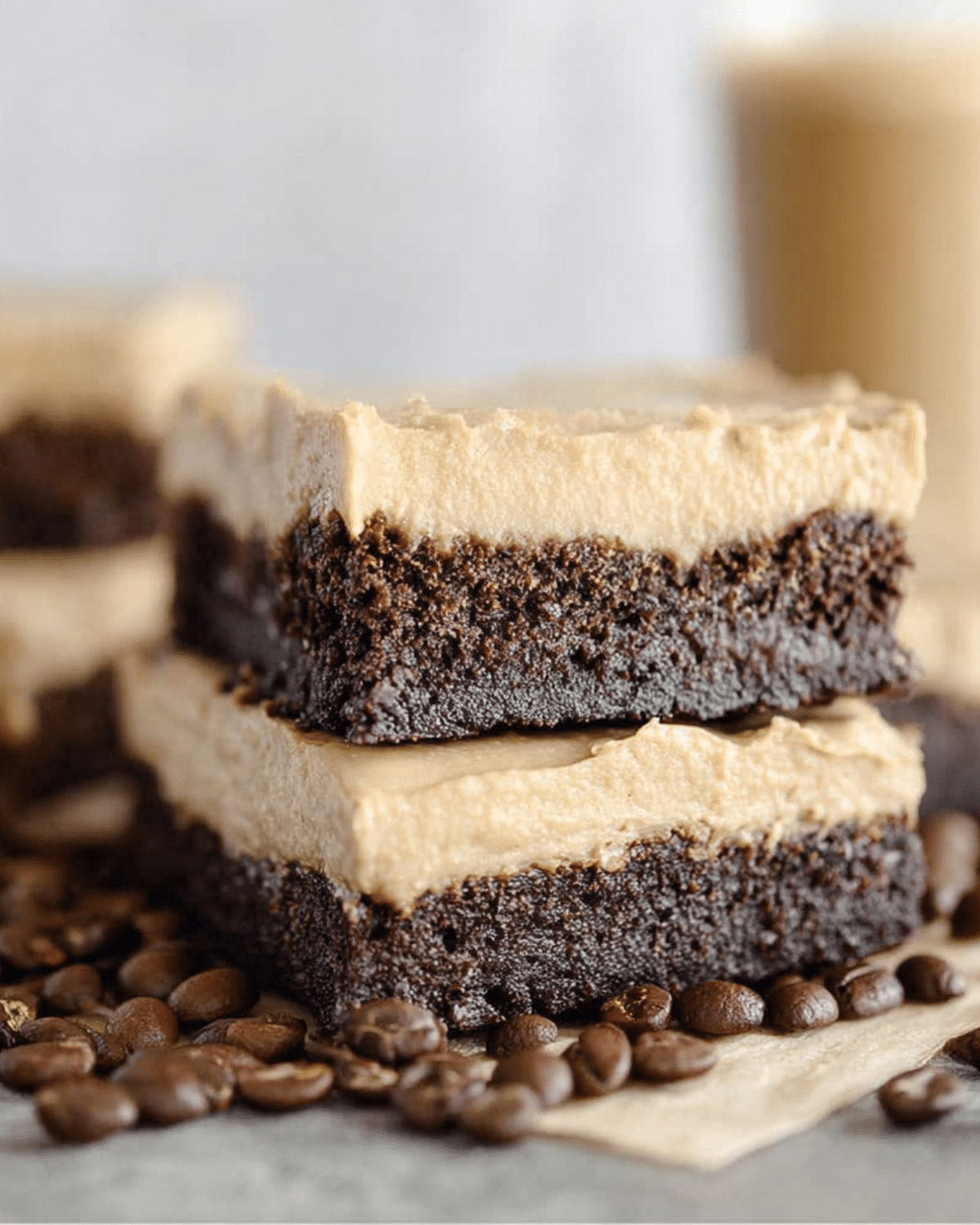 Two stacked frosted coffee brownies with a thick coffee cream layer on top of rich chocolate brownie, surrounded by coffee beans.