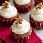 Gingerbread cupcakes with white frosting, each topped with a mini gingerbread cookie, in red cupcake liners.