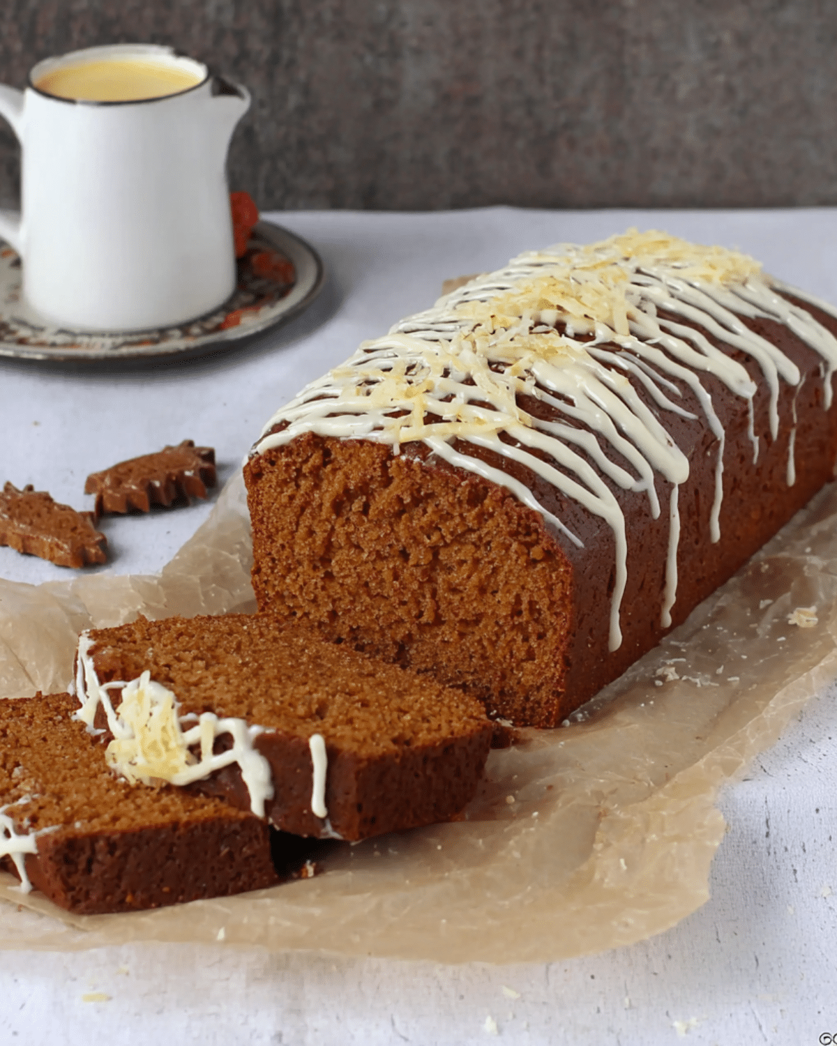 Sliced gingerbread loaf cake topped with white icing drizzle and grated cheese, served on parchment paper.