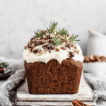 Gingerbread loaf with white frosting, topped with chopped pecans, cranberries, and rosemary sprigs, on a rustic white board.