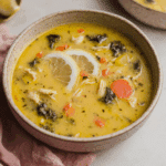 A bowl of Greek lemon chicken soup filled with shredded chicken, carrots, kale, and lemon slices in a creamy yellow broth.