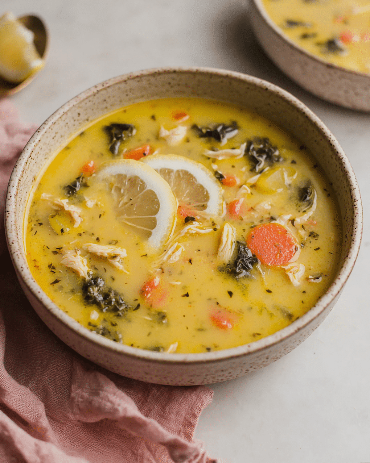 A bowl of Greek lemon chicken soup filled with shredded chicken, carrots, kale, and lemon slices in a creamy yellow broth.
