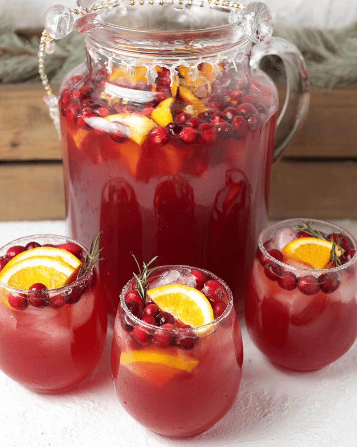 Festive holiday punch in a large glass pitcher and three glasses, garnished with orange slices, cranberries, and rosemary sprigs.