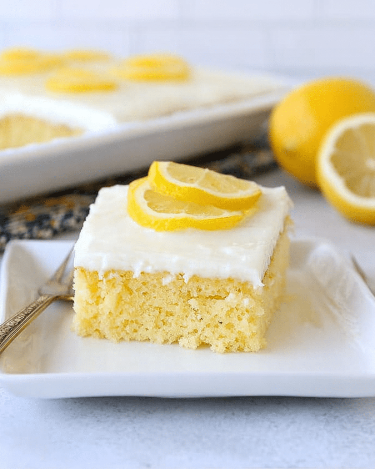 A slice of lemon sheet cake topped with white icing and garnished with lemon slices, served on a square white plate.