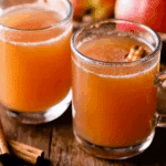 Two glass mugs of mulled cider with cinnamon sticks, surrounded by apples and spices on a wooden table.