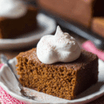 A square slice of old-fashioned gingerbread topped with a generous dollop of whipped cream and a sprinkle of cinnamon, served on a white plate.