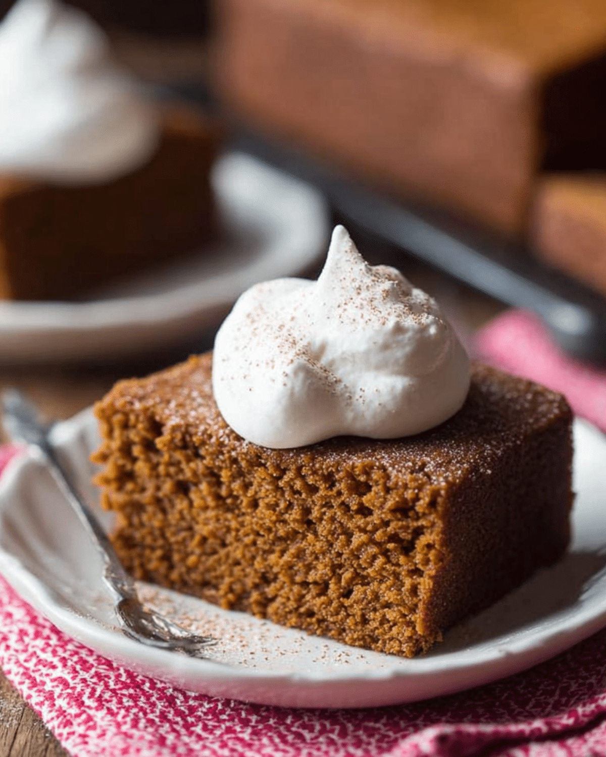 A square slice of old-fashioned gingerbread topped with a generous dollop of whipped cream and a sprinkle of cinnamon, served on a white plate.