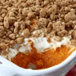A white casserole dish filled with old-fashioned sweet potato casserole, topped with a golden brown pecan crumble and a creamy layer, with a serving scooped out.