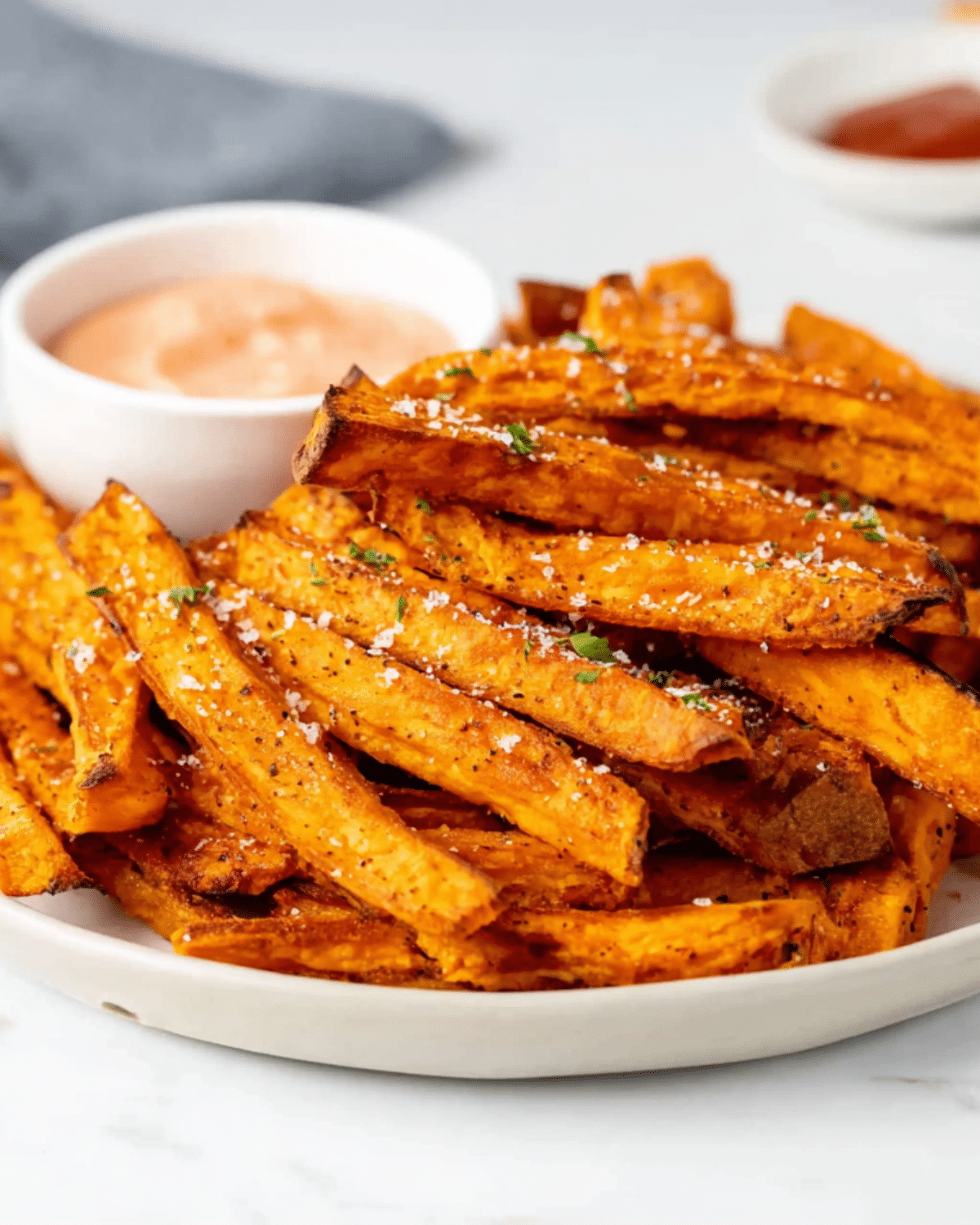 Crispy oven baked sweet potato fries on a white plate with a side of dipping sauce.