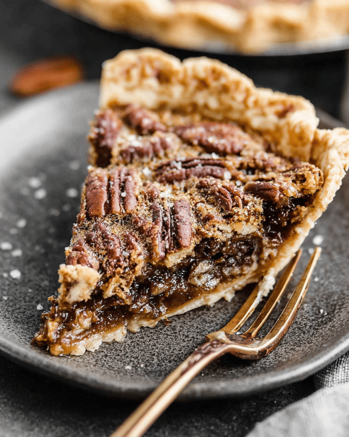 A slice of Pecan Pie with a flaky crust, gooey filling, and toasted pecans on top, served on a dark plate with a gold fork.