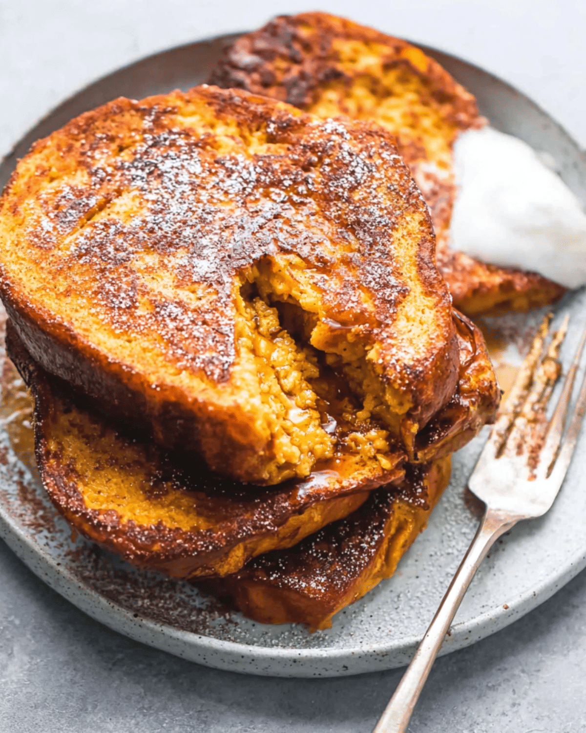 Stack of pumpkin French toast slices on a plate, topped with powdered sugar and served with whipped cream.