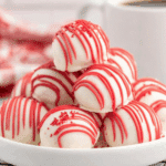 A stack of Red Velvet Cheesecake Bites covered in white chocolate and drizzled with red icing on a white plate.