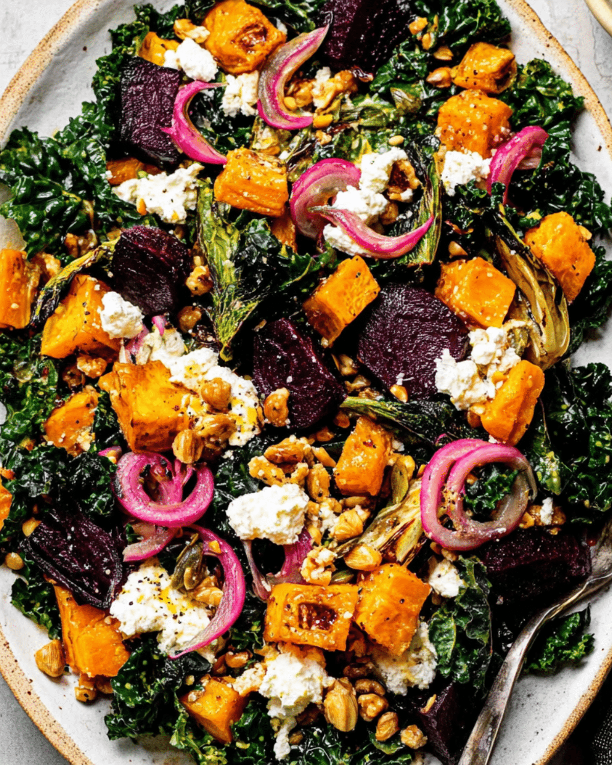 Colorful Roasted Vegetable Winter Salad with kale, beets, squash, pickled onions, goat cheese, and nuts on a white plate.