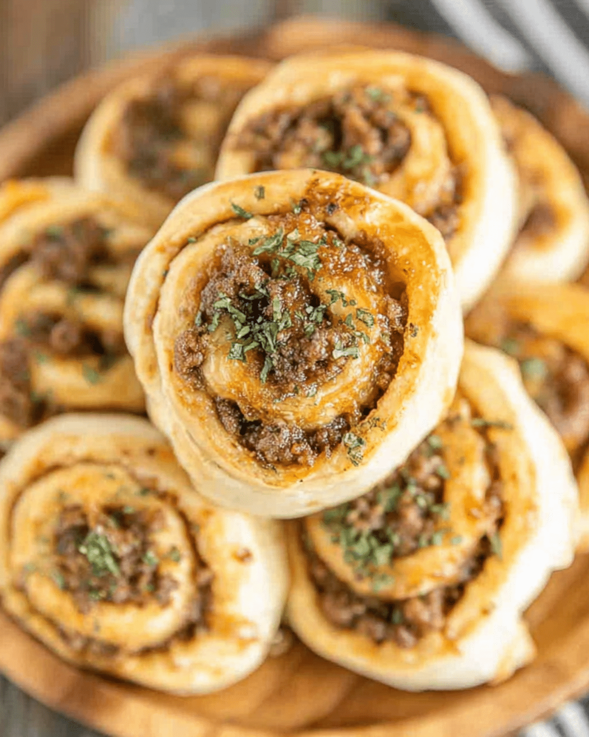 Sausage Pinwheels in Biscuit Dough stacked on a wooden plate, topped with herbs and featuring a golden, savory filling.