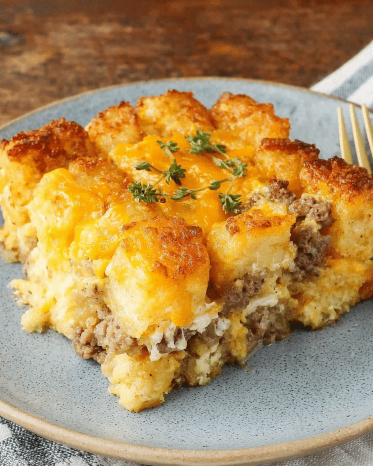 A serving of tater tot breakfast casserole with golden tater tots, melted cheddar cheese, sausage, and garnished with fresh herbs on a plate.