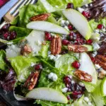 Winter Salad Recipe featuring mixed greens, sliced pears, pomegranate seeds, candied pecans, and crumbled cheese on a dark plate.