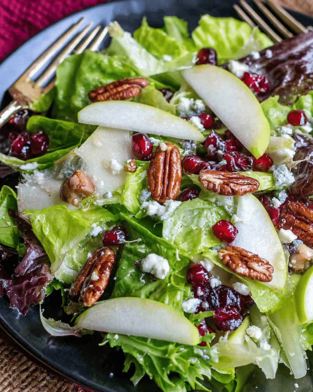 Winter Salad Recipe featuring mixed greens, sliced pears, pomegranate seeds, candied pecans, and crumbled cheese on a dark plate.