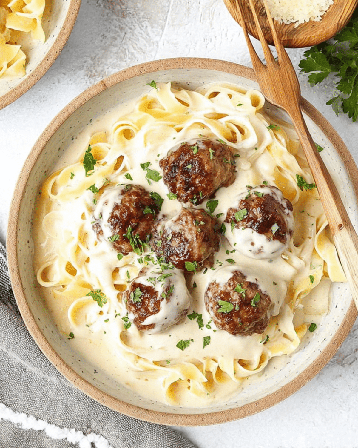 A bowl of Alfredo meatballs served over fettuccine pasta, topped with creamy Alfredo sauce and chopped parsley.