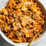 A bowl of beef noodle casserole with ground beef, egg noodles, melted cheese, and tomato sauce, topped with parsley.
