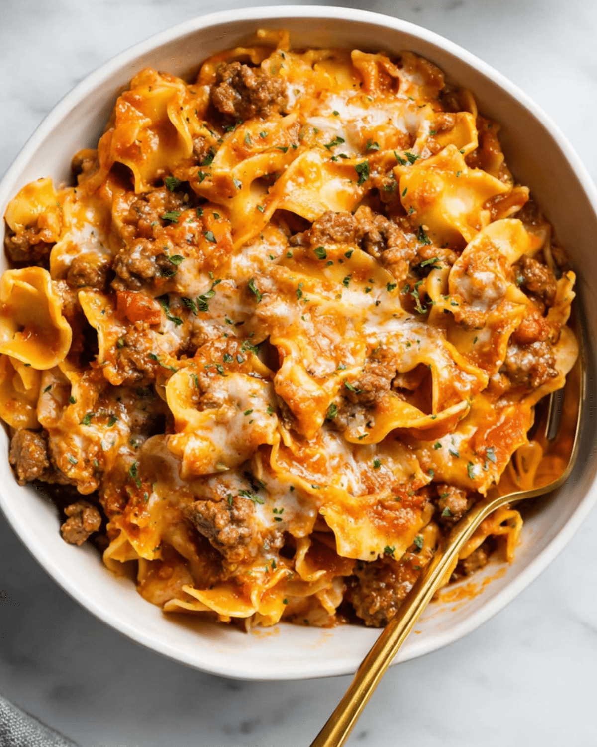 A bowl of beef noodle casserole with ground beef, egg noodles, melted cheese, and tomato sauce, topped with parsley.