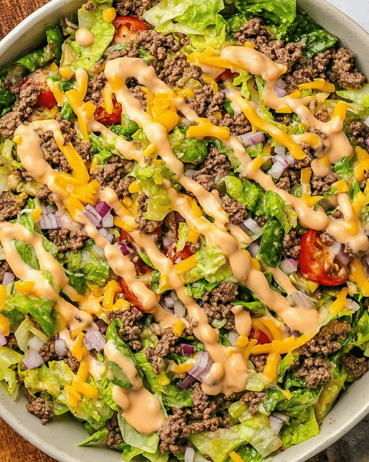 A bowl of Big Mac salad made with ground beef, shredded lettuce, cherry tomatoes, cheddar cheese, red onions, and drizzled with special sauce.