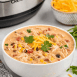 Bowl of creamy crack chicken chili topped with shredded cheddar cheese and cilantro, with visible corn, beans, and shredded chicken.