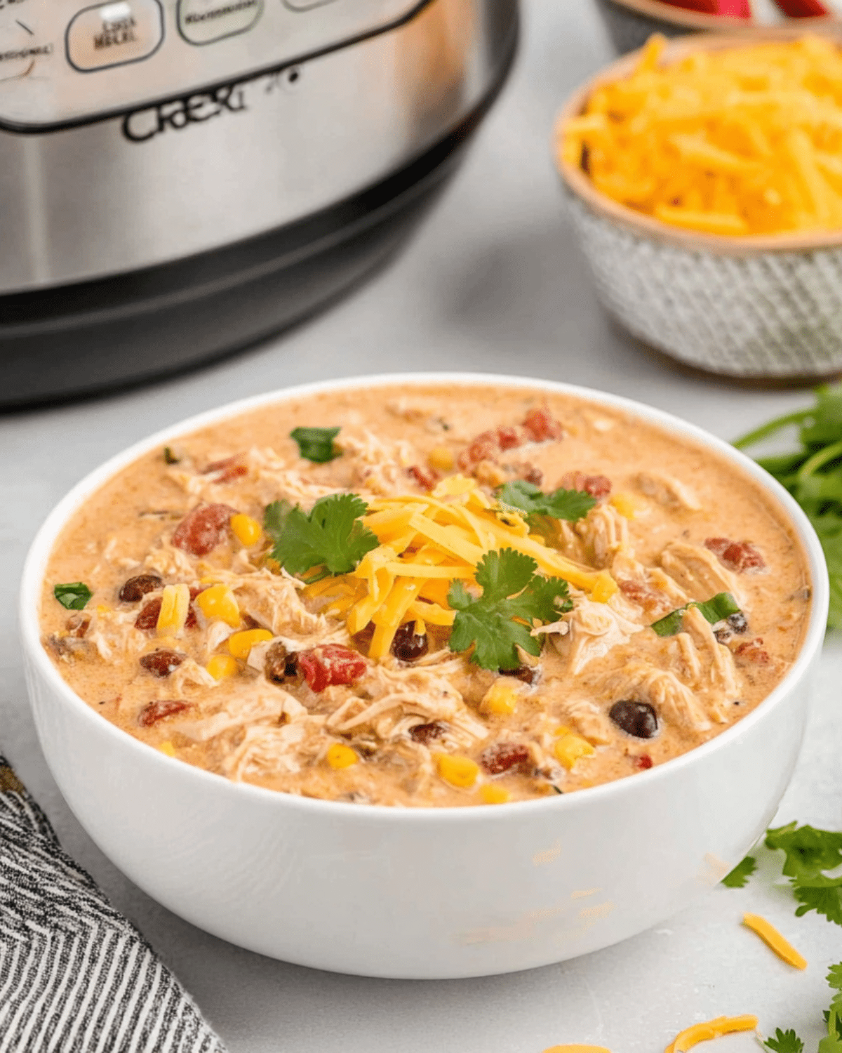 Bowl of creamy crack chicken chili topped with shredded cheddar cheese and cilantro, with visible corn, beans, and shredded chicken.
