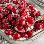 Glass dish filled with Cherries in the Snow, featuring bright red cherries in a glossy glaze over a fluffy white cream and cake layer, served with a spoon.