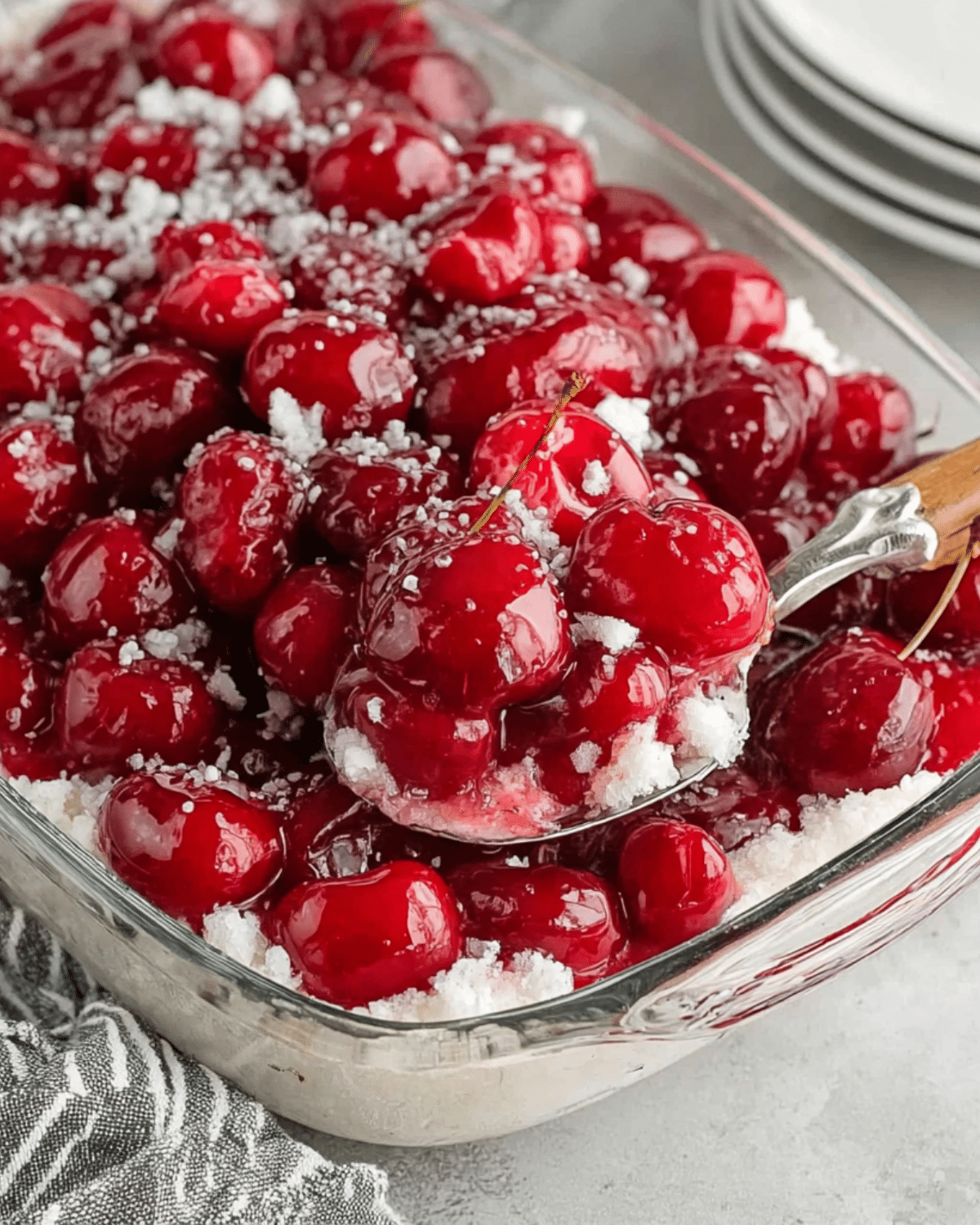 Glass dish filled with Cherries in the Snow, featuring bright red cherries in a glossy glaze over a fluffy white cream and cake layer, served with a spoon.