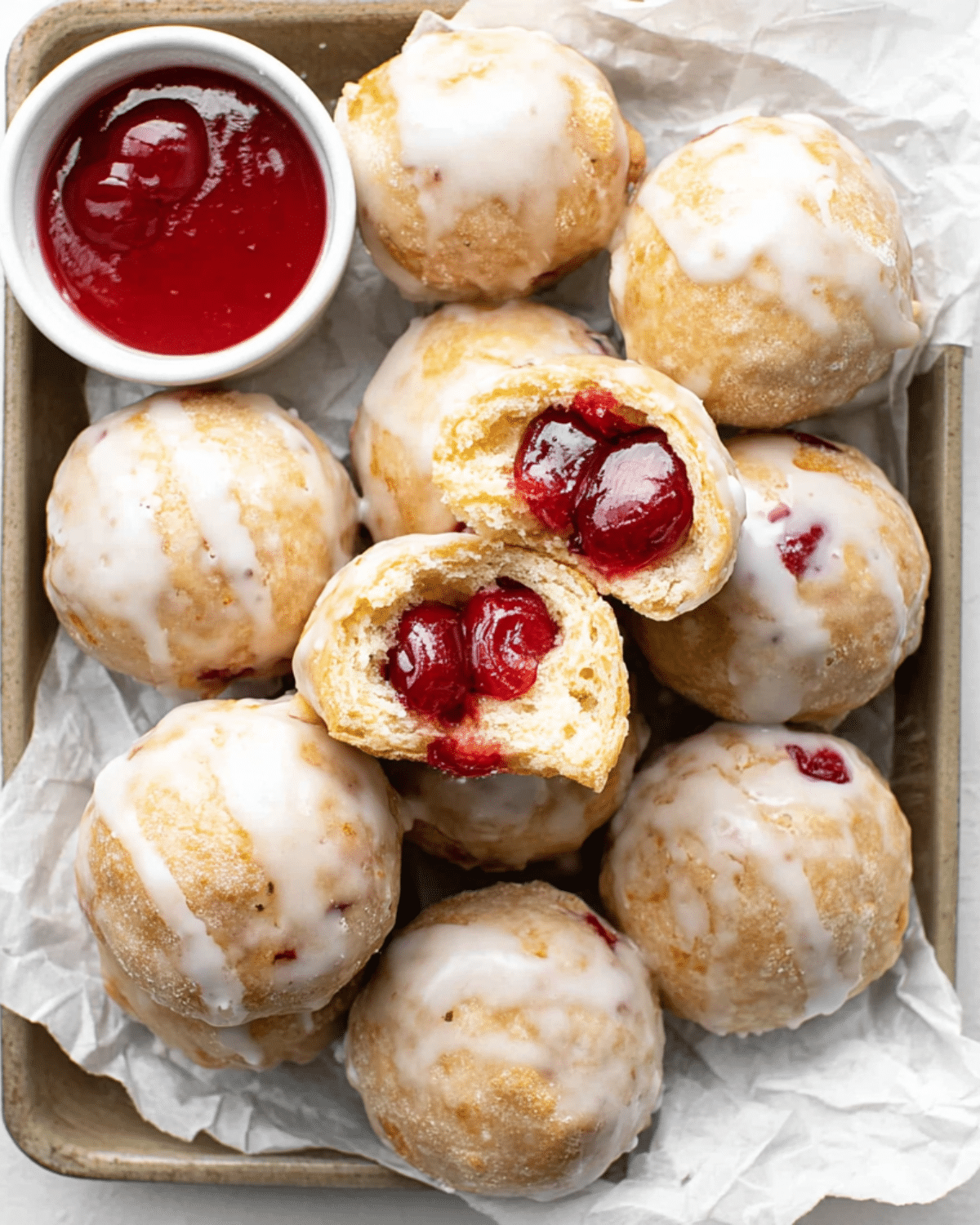 Tray of cherry pie bombs glazed with icing, with one opened to reveal a juicy cherry filling, served with a small bowl of cherry sauce.