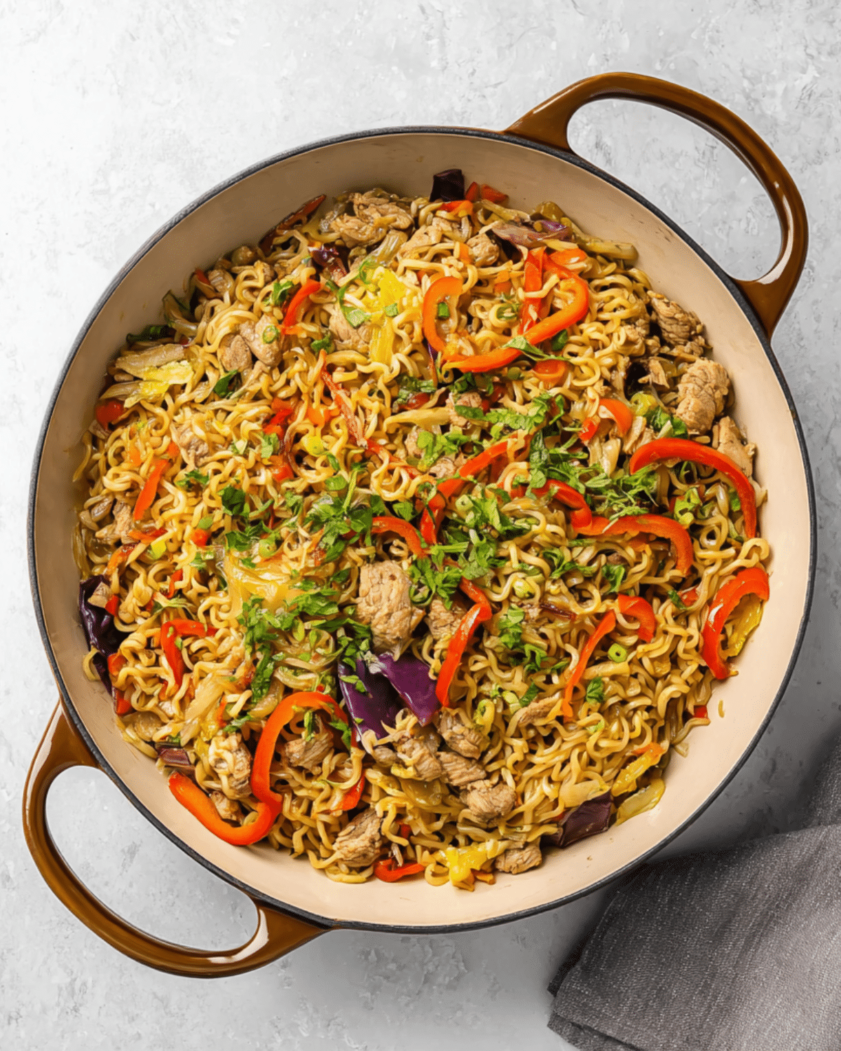 Pan filled with chicken ramen stir fry featuring noodles, sliced red bell peppers, cabbage, and chunks of chicken, garnished with chopped herbs.