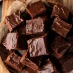 Rich and glossy chocolate fudge squares stacked in a wooden bowl lined with parchment paper, with a festive background.
