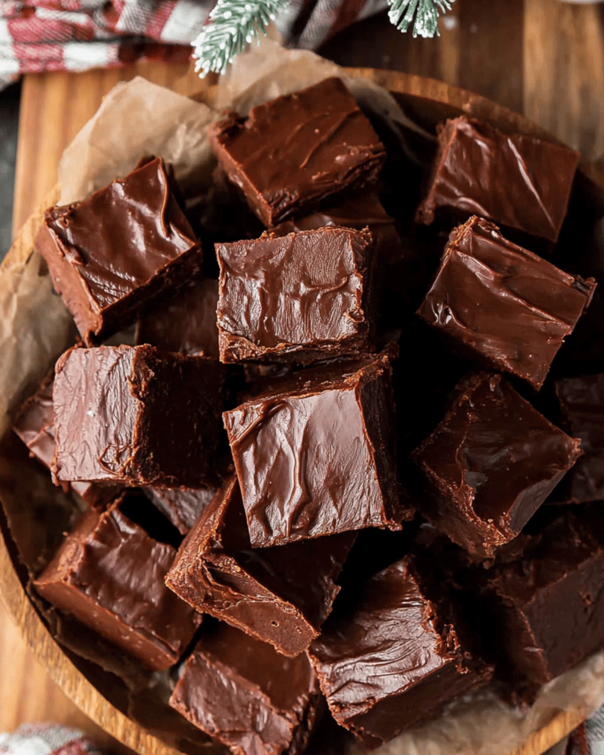 Rich and glossy chocolate fudge squares stacked in a wooden bowl lined with parchment paper, with a festive background.