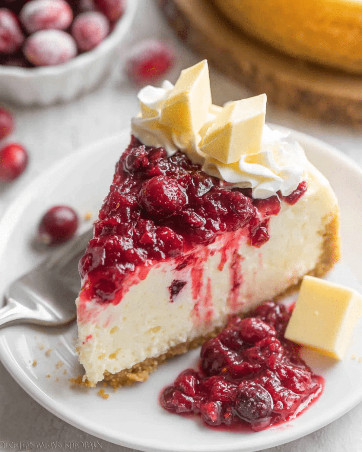 Slice of cranberry white chocolate cheesecake topped with cranberry sauce, whipped cream, and white chocolate pieces on a white plate.
