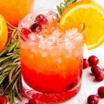 Glass of cranberry orange Christmas punch garnished with cranberries, orange slice, and rosemary sprig, surrounded by fresh cranberries.