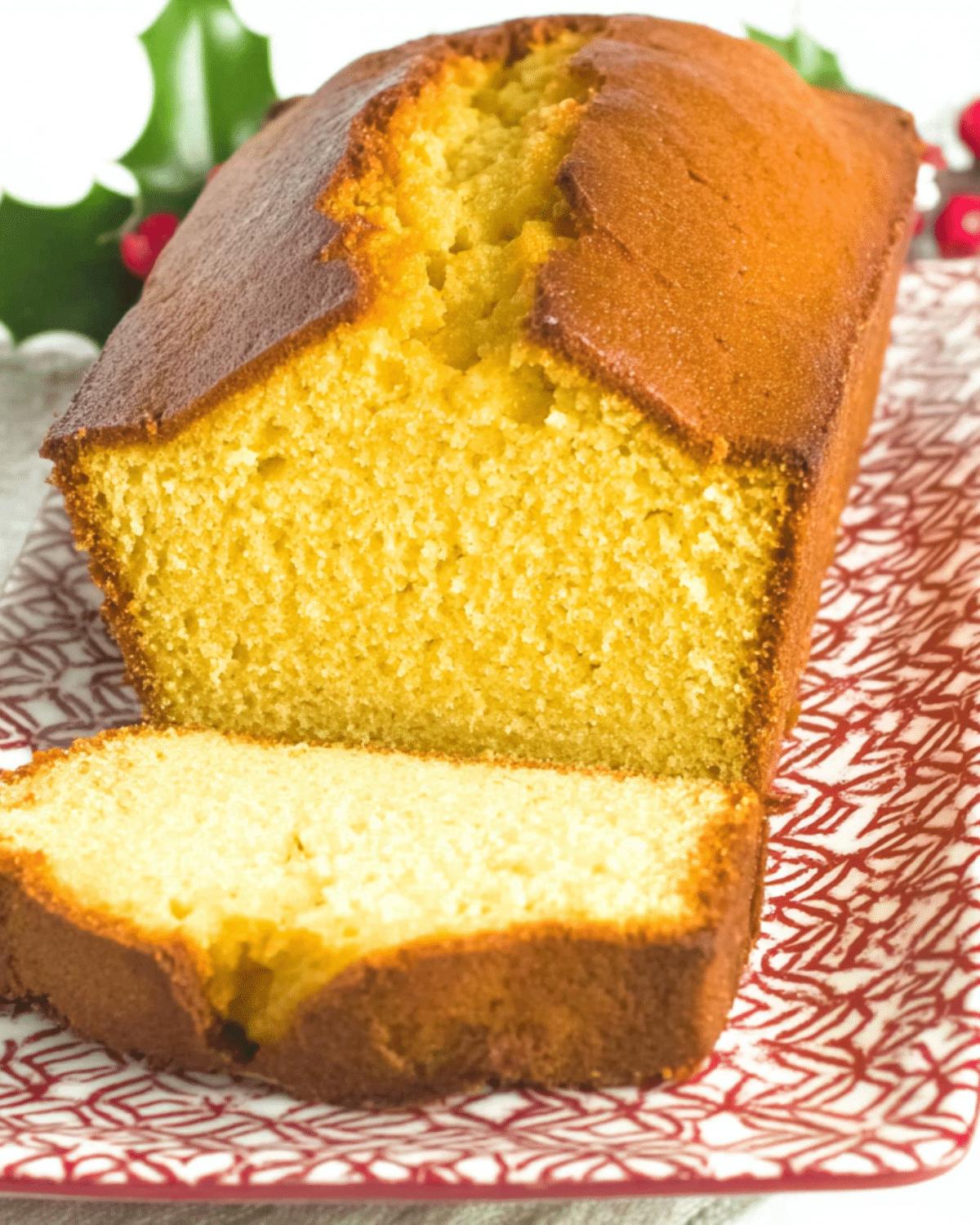Golden-brown eggnog bread loaf with a slice cut, showing its moist and fluffy interior, served on a festive red and white patterned plate.