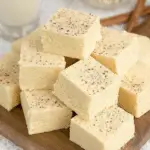 Creamy eggnog fudge squares topped with a sprinkle of nutmeg, stacked on a wooden board with cinnamon sticks and a glass of eggnog nearby.