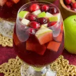 A glass of Fireball Sangria filled with red liquid, floating cranberries, and diced green and red apples.