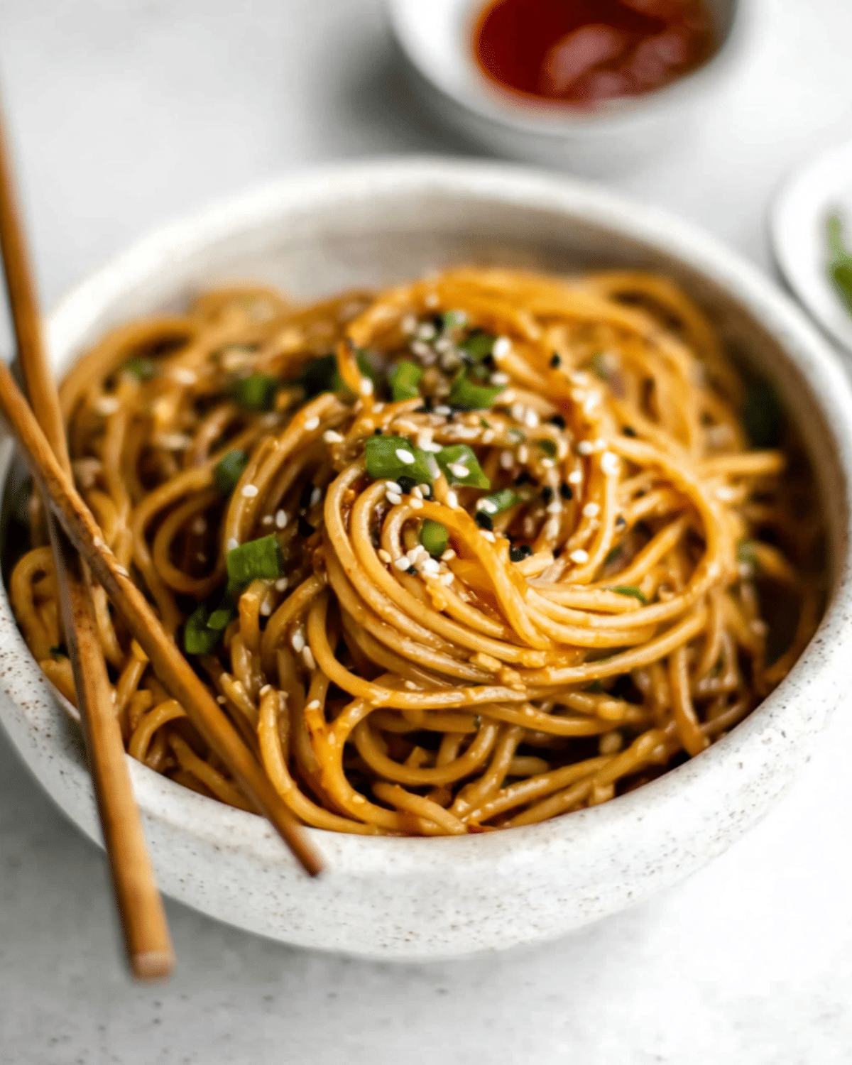 Bowl of garlic sesame noodles topped with sesame seeds and chopped green onions, served with chopsticks.
