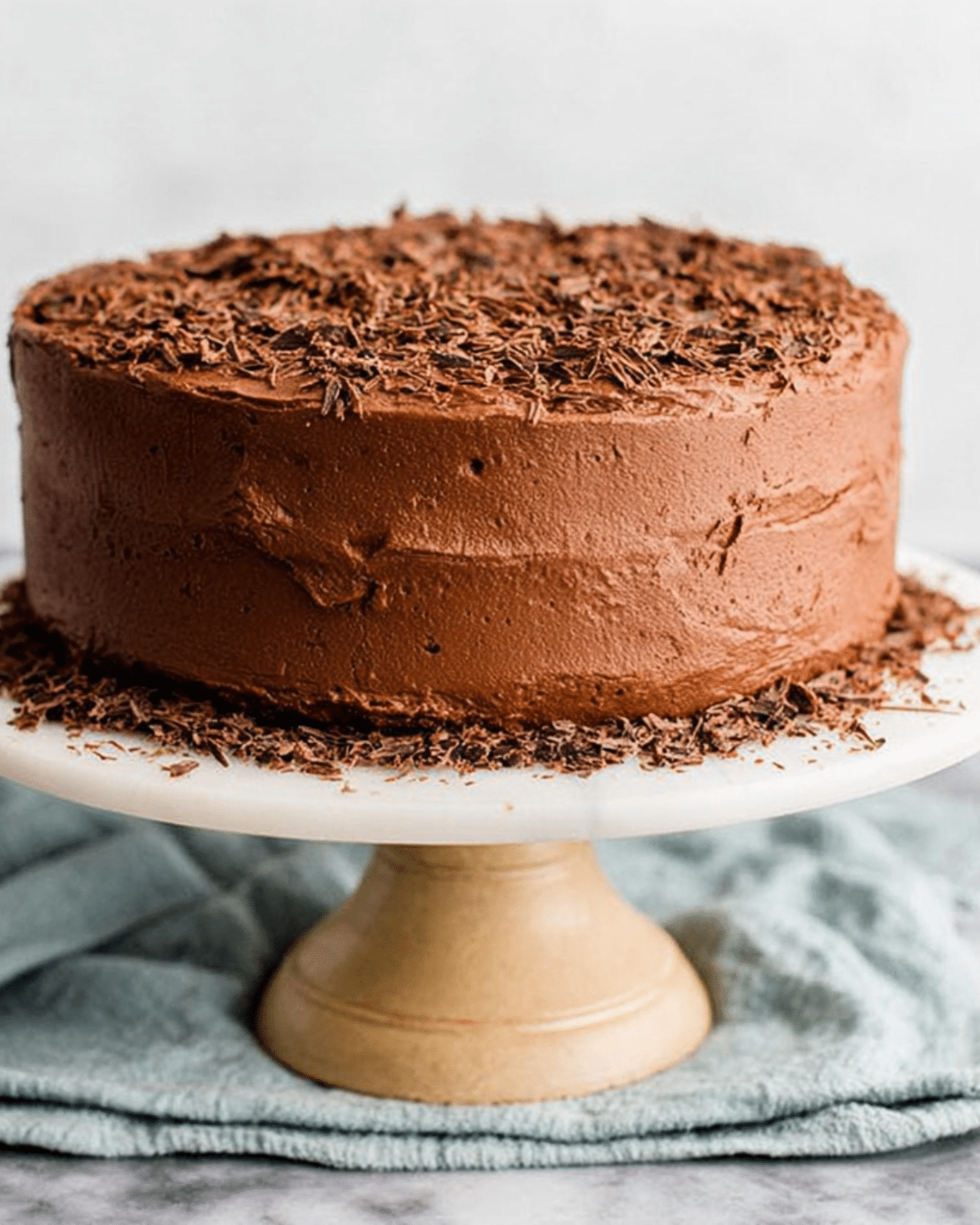 Rich and moist Hershey's Chocolate Cake covered in smooth chocolate frosting and topped with chocolate shavings, displayed on a cake stand.