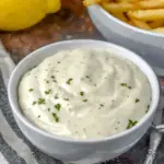 Bowl of homemade garlic aioli garnished with chopped herbs, placed next to a bowl of fries and fresh lemons.