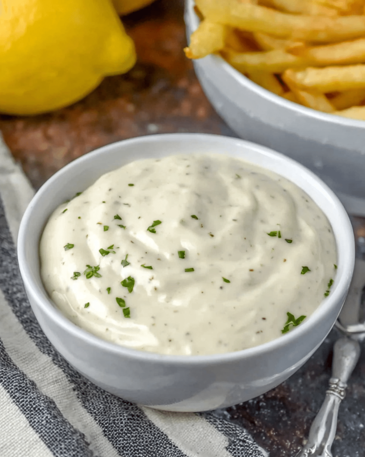 Bowl of homemade garlic aioli garnished with chopped herbs, placed next to a bowl of fries and fresh lemons.