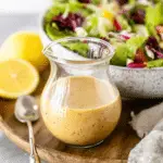 Glass jar of Lemon Vinaigrette Salad Dressing placed on a wooden tray with fresh lemon halves and a bowl of mixed salad in the background.